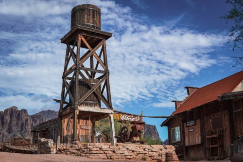 A rustic wooden water tower stands beside an old building in a desert landscape with mountains and blue skies.