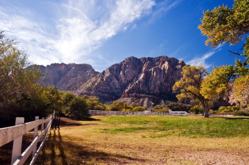 A scenic landscape featuring mountains, a blue sky, and a grassy field with a white fence and trees.