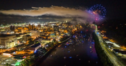 Aerial view of a city at night with fireworks, illuminated buildings, and boats on a river.