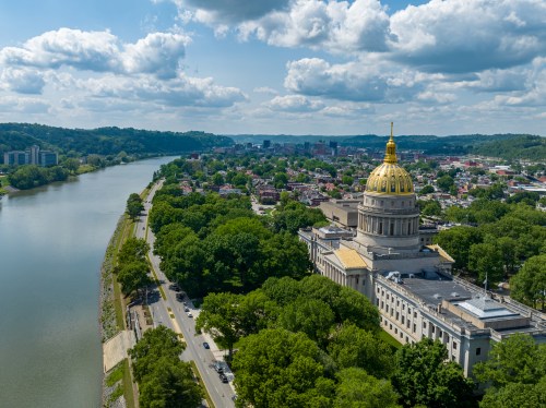 Aerial view of a river winding past a large building with a golden dome, surrounded by green trees and a city skyline.