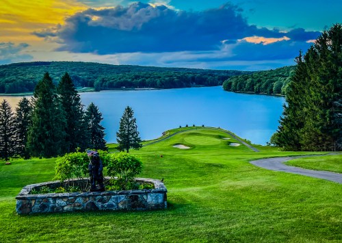 A scenic view of a golf course by a lake, surrounded by trees and a colorful sunset sky.