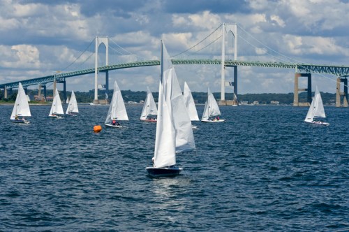 Sailboats with white sails on a calm waterway, with a large bridge in the background under a partly cloudy sky.