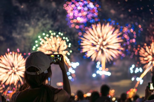 A person in a cap captures colorful fireworks on their camera against a night sky filled with vibrant bursts.
