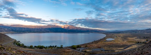 A panoramic view of a serene lake surrounded by mountains and a cloudy sky at dawn.