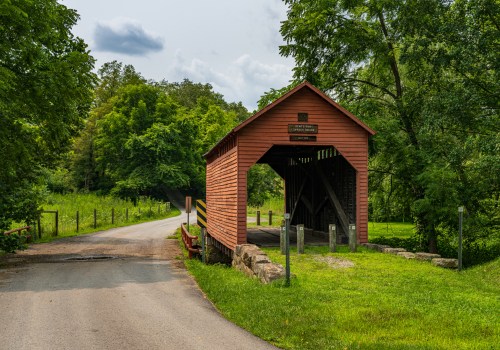 A red covered bridge stands at the edge of a grassy area, with a road leading into a wooded landscape.