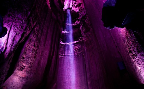 A vibrant purple-lit cave with a waterfall cascading down its rocky walls.