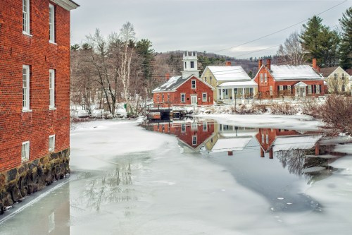 A snowy landscape featuring a calm river reflecting red brick buildings and trees, with a cloudy sky above.