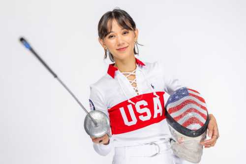 A young female fencer in a USA uniform holds a foil and a helmet with an American flag design, smiling confidently.