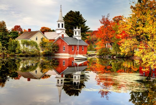A red church and colorful trees reflect in a calm lake, surrounded by autumn foliage and a serene landscape.