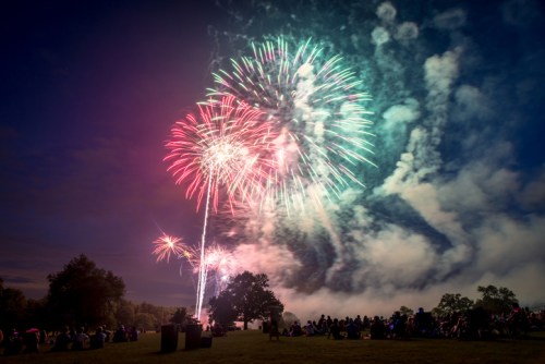 Colorful fireworks illuminate the night sky over a crowd gathered in a park, with smoke and trees in the foreground.