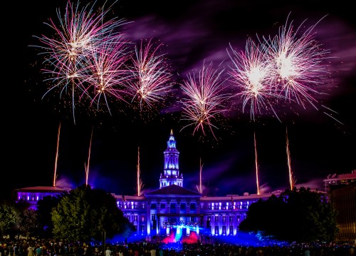 Colorful fireworks illuminate a historic building at night, with a crowd gathered below, celebrating a festive event.