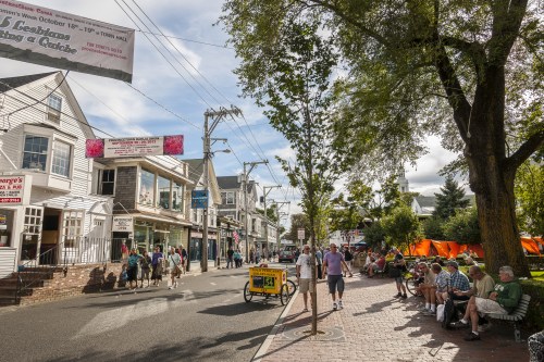 A busy street scene with shops, trees, and people sitting on benches, enjoying a sunny day.