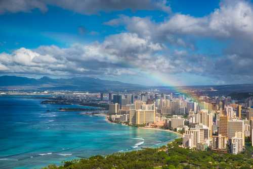 Aerial view of a vibrant city by the ocean, featuring skyscrapers, a rainbow, and lush greenery along the coastline.