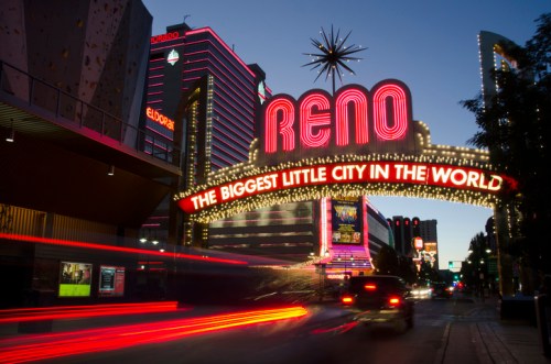 Neon sign reading "Reno: The Biggest Little City in the World" illuminated at dusk, with light trails from passing cars.