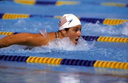 A swimmer in a white cap with an American flag competes in a butterfly stroke, splashing water in a pool.