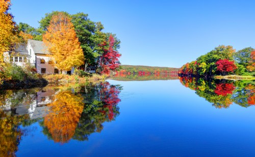 A serene lake reflects vibrant autumn foliage and a charming house under a clear blue sky.