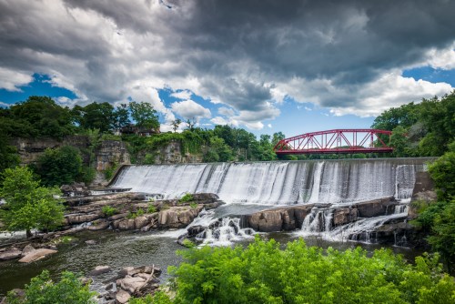 A scenic waterfall cascades over rocks, with a red bridge in the background under a cloudy sky. Lush greenery surrounds the area.
