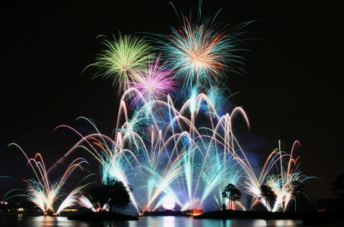 Colorful fireworks burst in the night sky over a lake, illuminating the scene with vibrant hues and reflections.