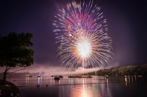 Colorful fireworks burst over a calm lake at night, reflecting in the water with trees and boats in the foreground.