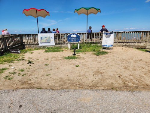 A sandy area with beach umbrellas and a sign for Main Street Beach, overlooking a calm sea and people in the background.