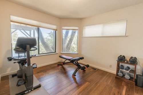 A bright home gym with a stationary bike, weight bench, dumbbells, and large windows letting in natural light.