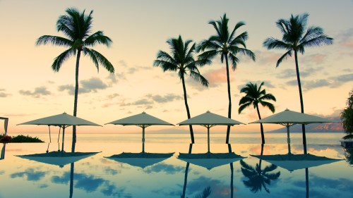 A serene sunset over a calm pool, with palm trees and shaded loungers reflecting in the water.