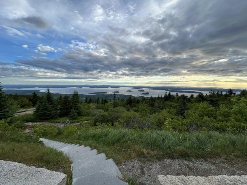 A scenic view from a hillside, featuring lush greenery, stone steps, and a cloudy sky over distant islands and water.