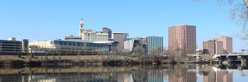 City skyline reflecting on a river, featuring modern buildings and a clear blue sky.