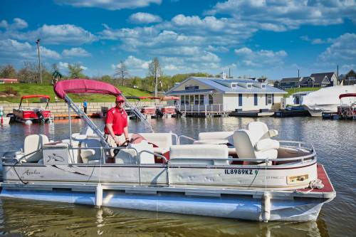 A person in a red shirt sits on a pontoon boat with a canopy, surrounded by water and a scenic background.
