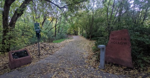 A wide, paved trail leads through a thick grove of trees in Salt Lake City on an autumn day. A sign next to the path says Hidden Hollow.
