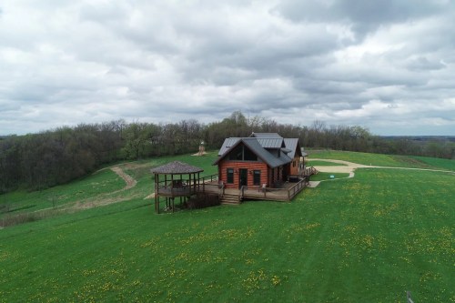 A rustic wooden house with a deck sits on a green lawn, surrounded by trees and under a cloudy sky.
