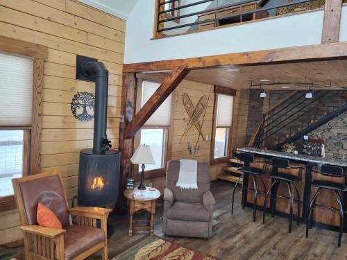 Cozy cabin interior with a wood stove, armchair, and a kitchen bar with stools, featuring rustic wooden walls and decor.