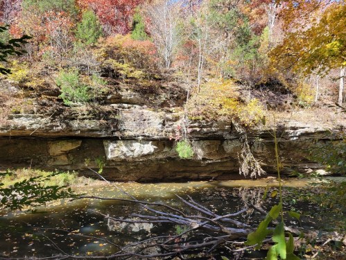 A serene landscape featuring a rocky cliff, colorful autumn foliage, and a calm pond reflecting the trees.