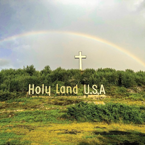 A large cross and the words "Holy Land U.S.A." are displayed under a rainbow in a green landscape.