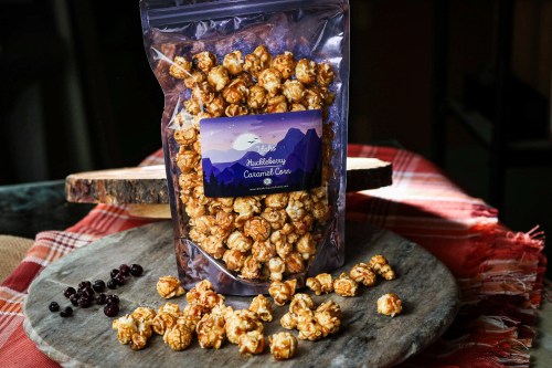 A bag of blueberry caramel popcorn on a wooden platter, with scattered popcorn and berries around it.
