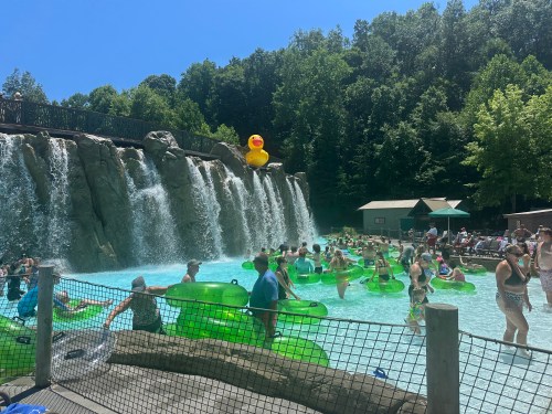 A busy water park scene with people floating in green tubes near a waterfall and a large rubber duck overhead.