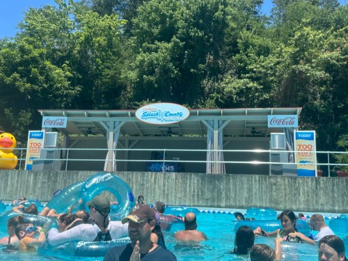 A crowded water park pool with people enjoying the water, a stage in the background, and trees surrounding the area.