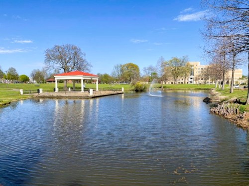 A serene park scene featuring a pond, gazebo, and fountain under a clear blue sky. Trees line the grassy shore.