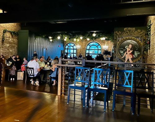 A lively restaurant scene with diners at tables, exposed brick walls, and a mural of a woman on the wall.