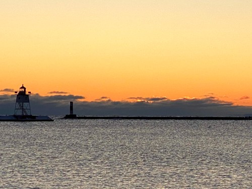 A lighthouse stands by the water at sunset, with a colorful sky and distant clouds reflecting on the calm sea.