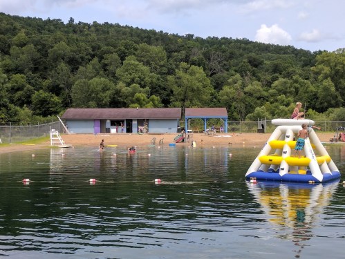 A lake scene with children playing on an inflatable structure, a sandy beach, and trees in the background.