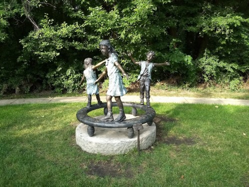 A bronze sculpture of three children playing on a circular platform, surrounded by greenery.