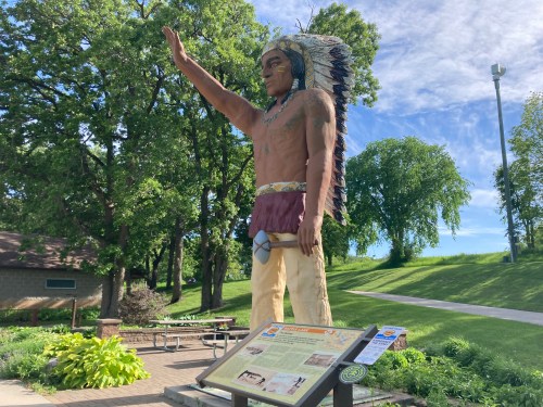 A large statue of a Native American chief with a feathered headdress, standing in a park with trees and a sign nearby.