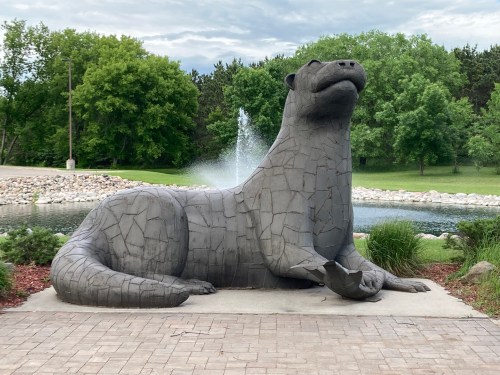 A large stone sculpture of an otter sits by a pond, surrounded by greenery and a fountain in the background.