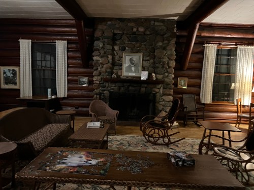 Cozy log cabin interior with a stone fireplace, rocking chairs, and a puzzle on the table, illuminated by soft lighting.