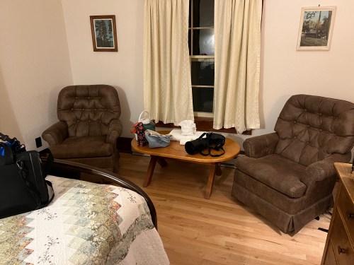 A cozy room with two brown armchairs, a wooden table, and a bed with a quilt, featuring a window with curtains.