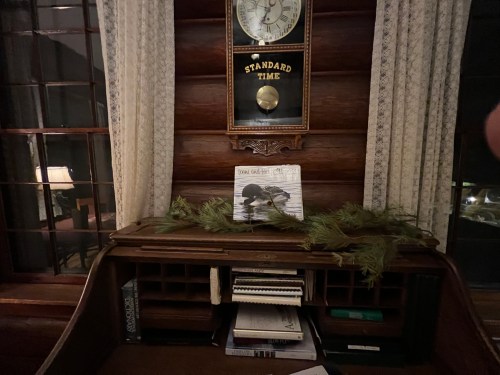 A wooden desk with a clock, a decorative print, and pine branches, set against a log cabin wall.