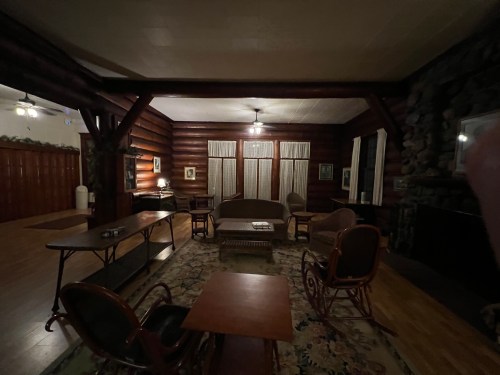 Cozy log cabin interior with wooden beams, a stone fireplace, and rustic furniture arranged around a patterned rug.