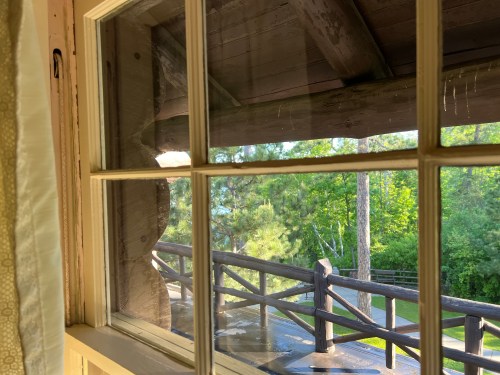View through a window showing wooden beams and a balcony overlooking a green landscape.