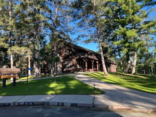 A rustic lodge surrounded by tall trees, with a pathway leading to the entrance and a sign for Douglas Lodge Restaurant.